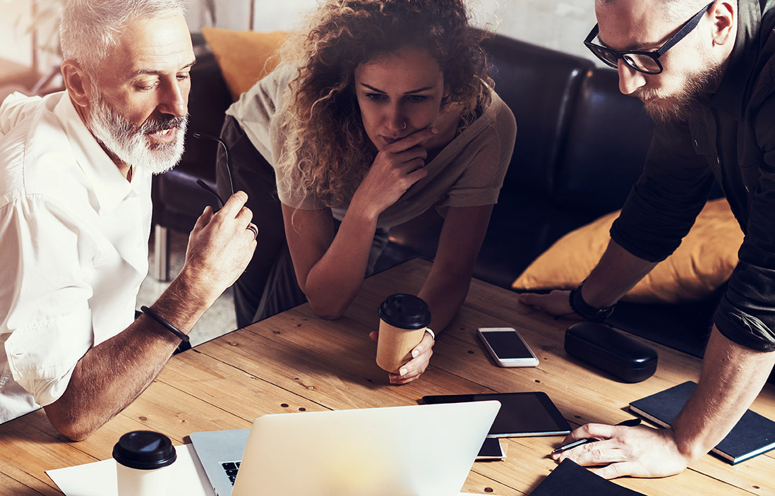 Group of three professionals discussing strategies over a laptop, with coffee cups and smartphones on a wooden table, reflecting collaboration in healthcare consulting for the HRSA 340B program.